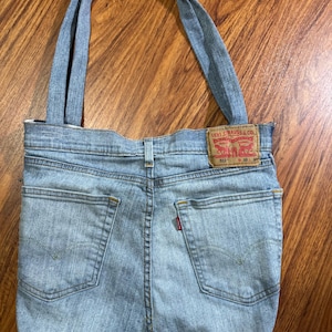 Op de afbeelding: Een lichtblauwe denim tote bag gemaakt van hergebruikte jeans. De tas heeft de achterzakken en het label van de jeans, met een rood Levi Strauss & Co. label. De tas heeft twee denim handvatten.
