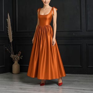 Puede incluir: Un vestido de satén naranja con escote cuadrado y detalles de lazo en los hombros. El vestido largo tiene cintura fruncida y falda fluida. La persona lleva tacones rojos.