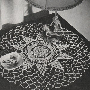 May include: A white crocheted doily with a delicate lace pattern. The doily is placed on a dark surface and features a central circular design with intricate details.