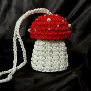 May include: A handmade crochet mushroom with a red cap dotted with white beads and a white stem. A white cord is attached to the top for hanging. The mushroom is set against a dark background.