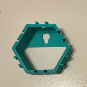 K&ouml;nnte beinhalten: Ein t&uuml;rkisfarbenes, sechseckiges Wandregal mit einer schl&uuml;ssellochf&ouml;rmigen &Ouml;ffnung zum Aufh&auml;ngen. Das Regal hat ein geometrisches Design mit mehreren kleinen, rechteckigen Kerben an den Seiten.
