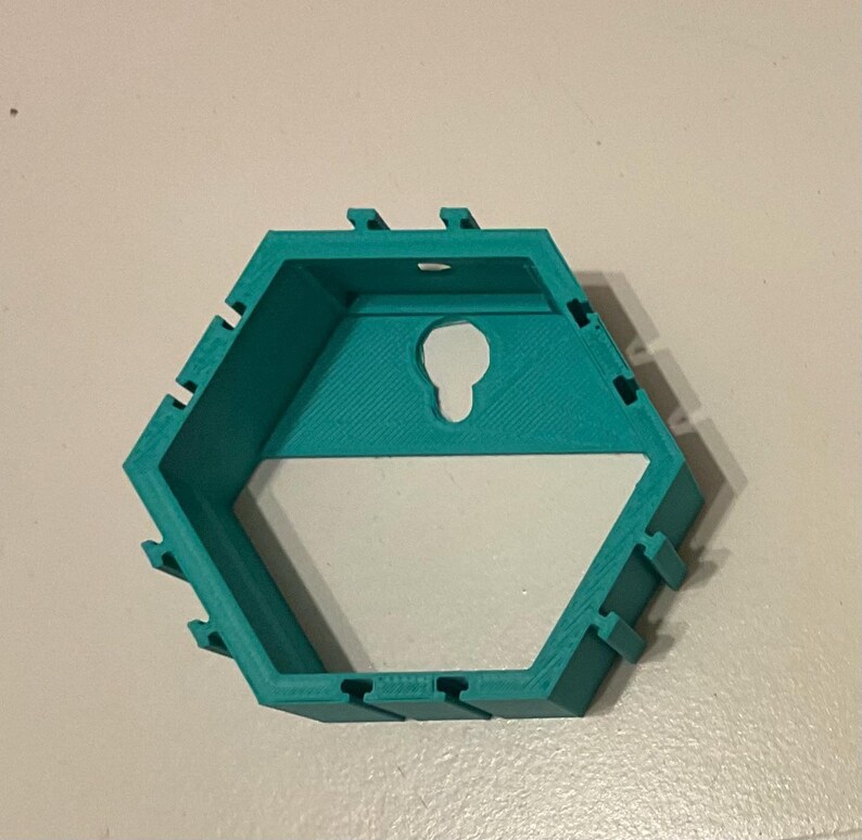 K&ouml;nnte beinhalten: Ein t&uuml;rkisfarbenes, sechseckiges Wandregal mit einer schl&uuml;ssellochf&ouml;rmigen &Ouml;ffnung zum Aufh&auml;ngen. Das Regal hat ein geometrisches Design mit mehreren kleinen, rechteckigen Kerben an den Seiten.