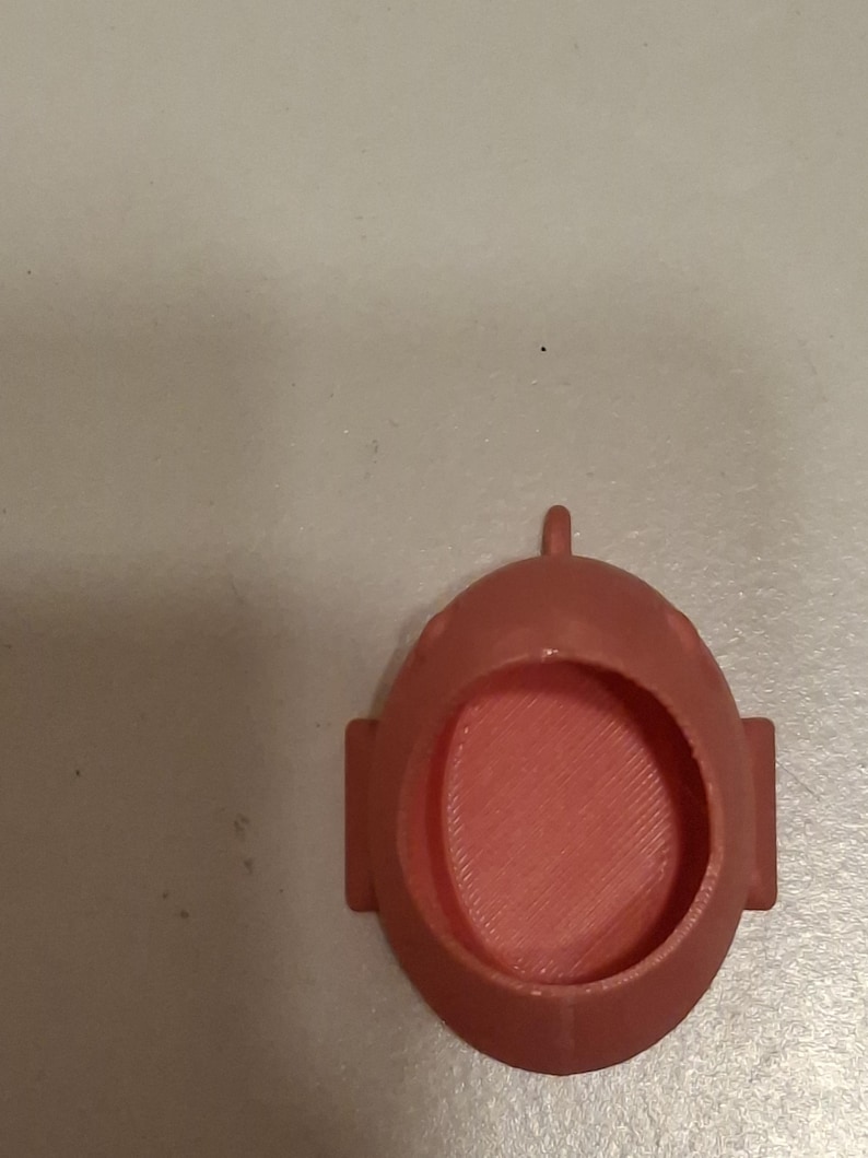 May include: A coral-colored, 3D-printed object with an oval shape and a circular opening. It has a small loop at the top and two rectangular protrusions on the sides. The interior has a textured, ridged pattern.