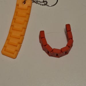 Puede incluir: Eslabones de cadena de pl&aacute;stico naranja y rojo. Una cadena naranja est&aacute; unida a un llavero con un anillo plateado. La otra cadena es roja y tiene forma de U. Las cadenas est&aacute;n sobre una superficie blanca.
