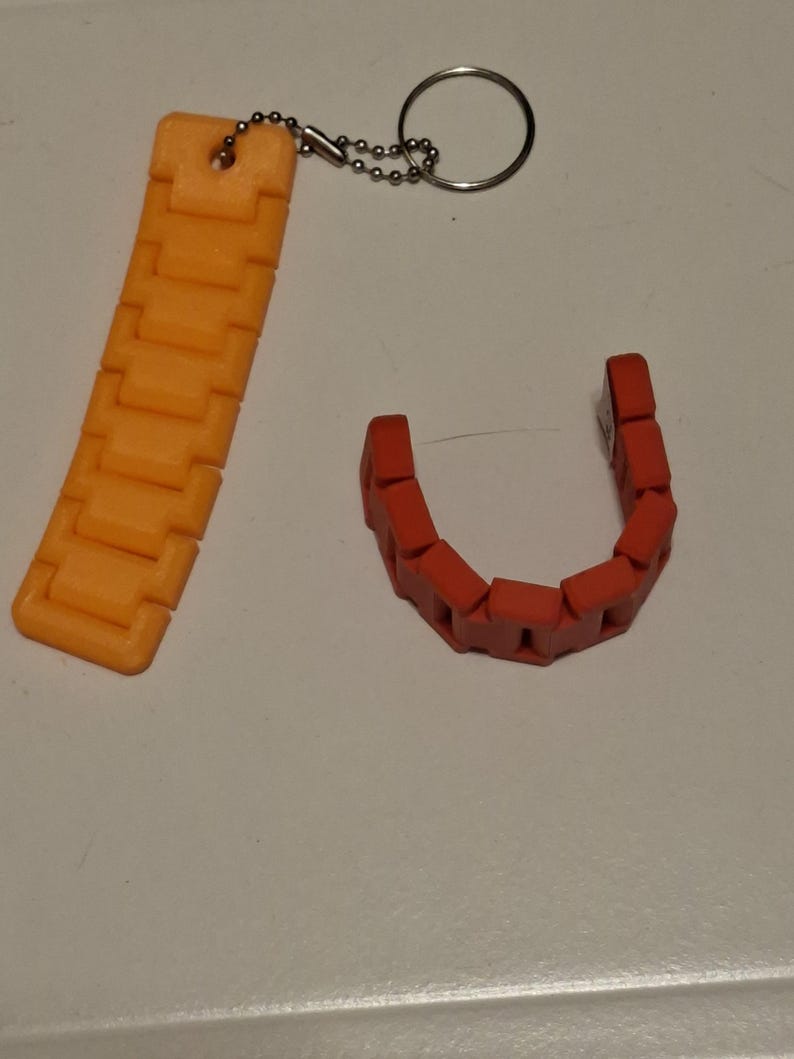 Puede incluir: Eslabones de cadena de pl&aacute;stico naranja y rojo. Una cadena naranja est&aacute; unida a un llavero con un anillo plateado. La otra cadena es roja y tiene forma de U. Las cadenas est&aacute;n sobre una superficie blanca.