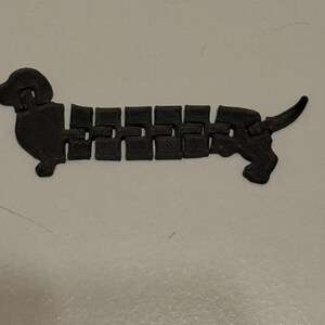 May include: A black articulated dachshund dog toy. The toy is made of connected rectangular segments, allowing it to bend and move. The dog has a head, tail, and four small feet. The toy is on a white surface.
