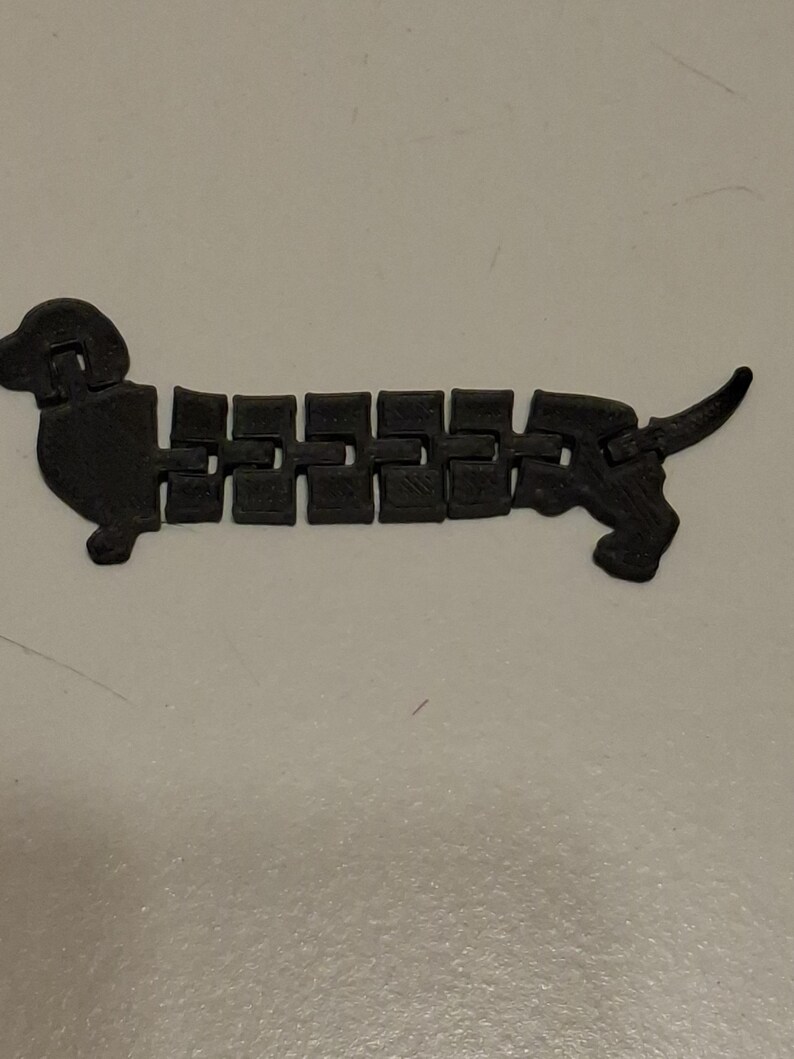 May include: A black articulated dachshund dog toy. The toy is made of connected rectangular segments, allowing it to bend and move. The dog has a head, tail, and four small feet. The toy is on a white surface.