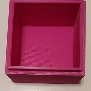 May include: A magenta-colored, square-shaped container with a textured interior and a raised lip. The box is made of a solid material and has a simple, geometric design. The container is empty and ready for use.