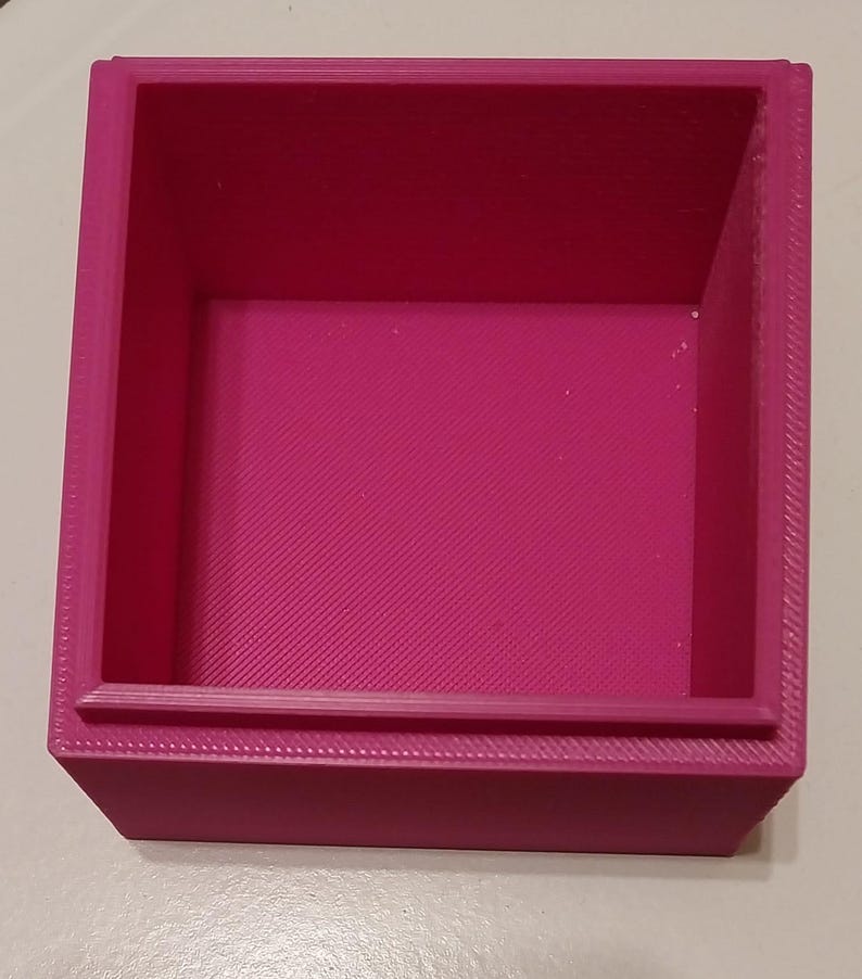 May include: A magenta-colored, square-shaped container with a textured interior and a raised lip. The box is made of a solid material and has a simple, geometric design. The container is empty and ready for use.