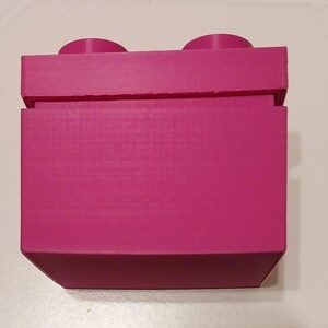 May include: A magenta-colored building block toy with two circular studs on top. The block has a rectangular shape with a textured surface and is made of plastic. The toy is designed to connect with other blocks.