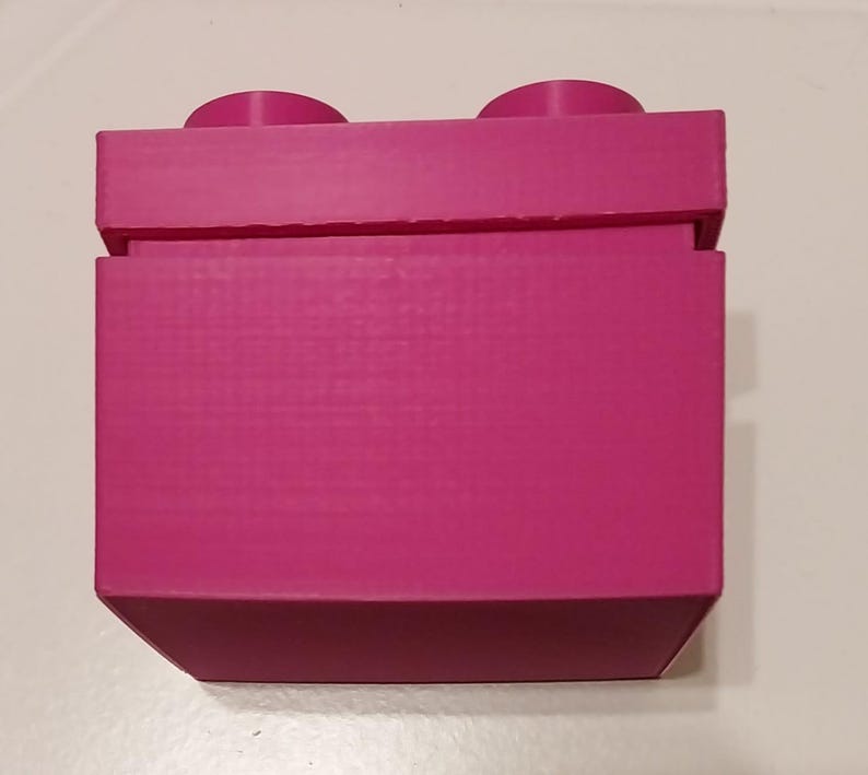 May include: A magenta-colored building block toy with two circular studs on top. The block has a rectangular shape with a textured surface and is made of plastic. The toy is designed to connect with other blocks.
