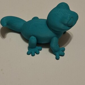 Op de afbeelding: Een turquoise 3D-geprinte hagedissenfiguur. De hagedis heeft een afgerond lichaam, een lange staart en vier kleine voeten. De figuur is gemaakt van een solide materiaal en heeft een getextureerd oppervlak.