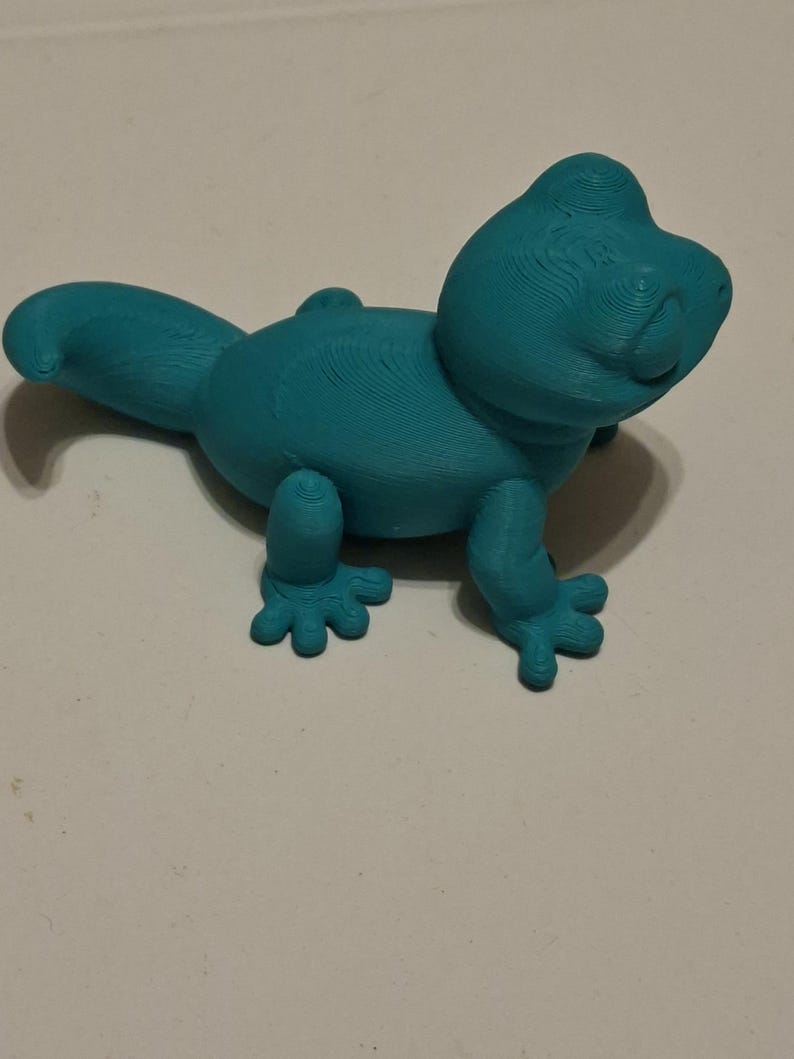 Op de afbeelding: Een turquoise 3D-geprinte hagedissenfiguur. De hagedis heeft een afgerond lichaam, een lange staart en vier kleine voeten. De figuur is gemaakt van een solide materiaal en heeft een getextureerd oppervlak.