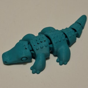 Op de afbeelding: Een turquoise gearticuleerd alligator speelgoed. Het plastic speelgoed heeft een gesegmenteerd lichaam voor flexibele bewegingen. De alligator heeft een gedetailleerde kop, poten en staart, met kleine bultjes op zijn rug. Een leuk, tactiel speelgoed.
