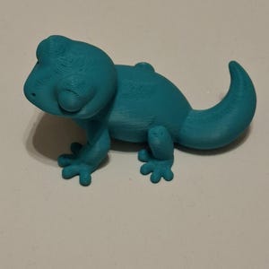 Op de afbeelding: Een turquoise 3D-geprint gekko-beeldje. Het reptiel heeft een afgeronde kop, kleine pootjes en een gekrulde staart. Het beeldje is gemaakt van een stevig materiaal en staat op een wit oppervlak.