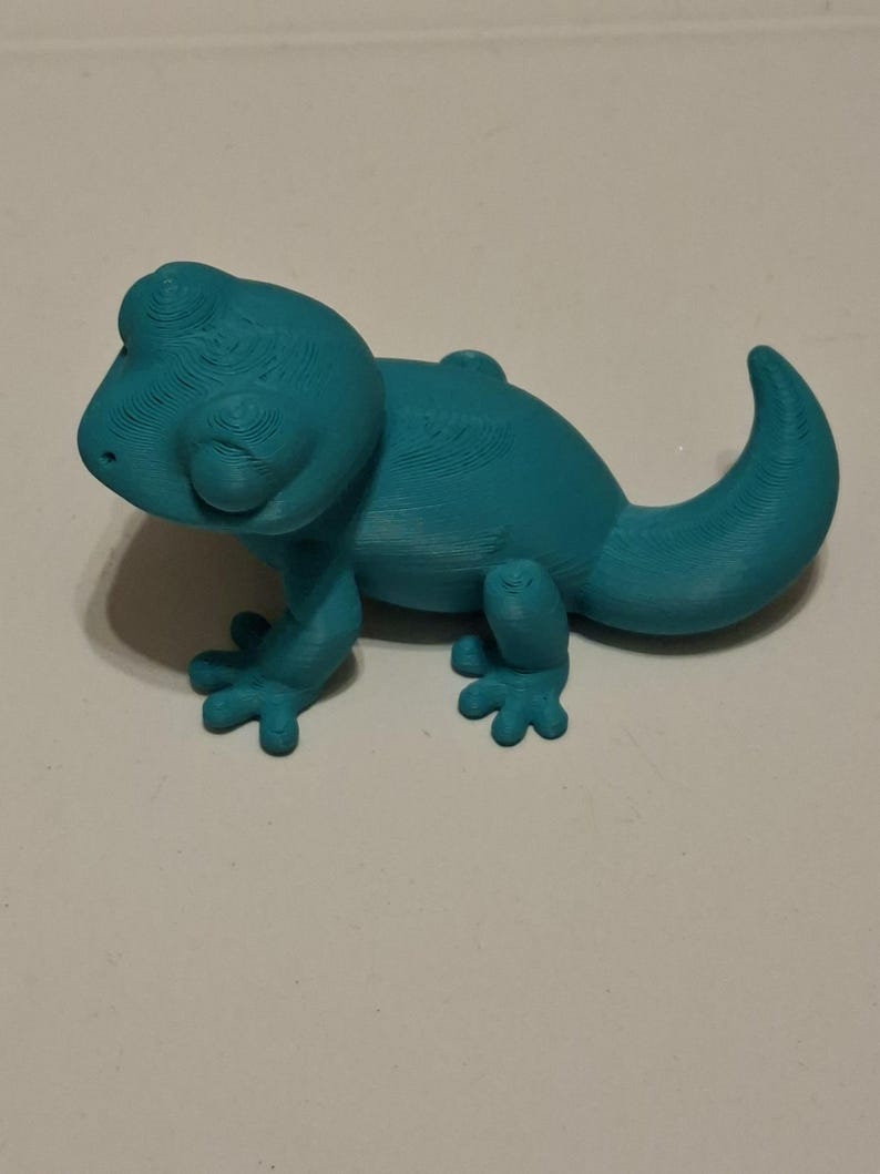 Op de afbeelding: Een turquoise 3D-geprint gekko-beeldje. Het reptiel heeft een afgeronde kop, kleine pootjes en een gekrulde staart. Het beeldje is gemaakt van een stevig materiaal en staat op een wit oppervlak.