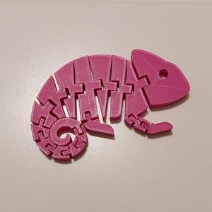 May include: A bright pink, articulated chameleon figurine. The toy is 3D-printed and features a segmented body, allowing for flexible posing. The chameleon has a curled tail and a detailed head with eye sockets.