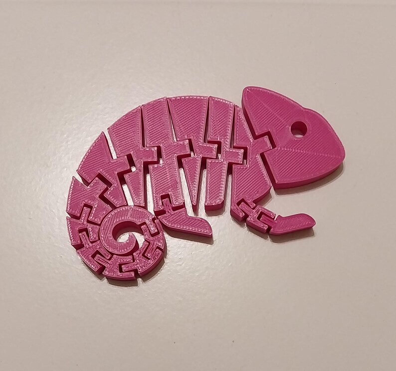 May include: A bright pink, articulated chameleon figurine. The toy is 3D-printed and features a segmented body, allowing for flexible posing. The chameleon has a curled tail and a detailed head with eye sockets.