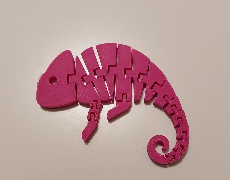 May include: A bright pink, articulated chameleon toy. The toy is made of connected segments, allowing it to be posed. The chameleon has a curled tail and a small hole for an eye. The toy is designed to be a fun and interactive item.