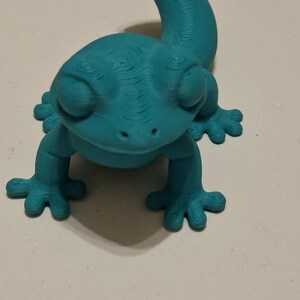 Op de afbeelding: Een turquoise 3D-geprinte gekko-figuur. De hagedis heeft een gebogen staart, vier kleine voetjes en een gedetailleerd, getextureerd oppervlak. De gekko is effen van kleur en lijkt van plastic te zijn.