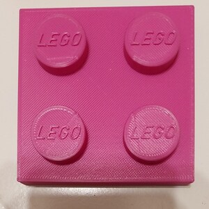 May include: A bright pink LEGO brick with four raised studs, each bearing the LEGO logo. The block is square and has a textured surface. A classic toy building block.