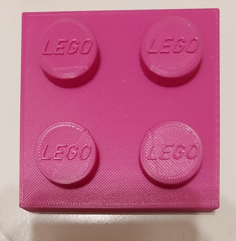May include: A bright pink LEGO brick with four raised studs, each bearing the LEGO logo. The block is square and has a textured surface. A classic toy building block.