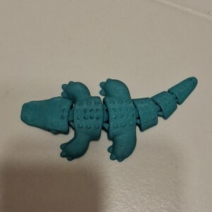 Op de afbeelding: Een turquoise gearticuleerd dinosaurus speelgoed. Het speelgoed is gesegmenteerd, waardoor flexibele beweging mogelijk is. De dinosaurus heeft een gedetailleerde textuur en is ontworpen om op een gestileerde reptiel te lijken. Het speelgoed is gemaakt van plastic.