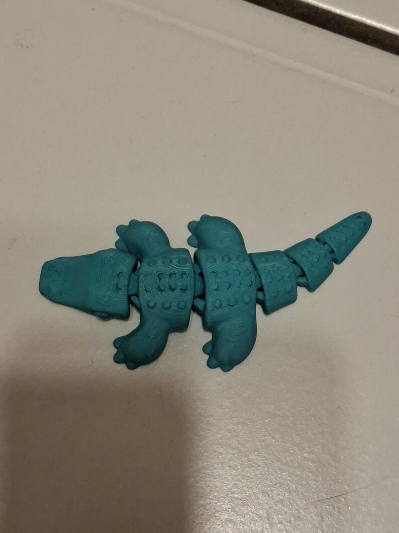 Op de afbeelding: Een turquoise gearticuleerd dinosaurus speelgoed. Het speelgoed is gesegmenteerd, waardoor flexibele beweging mogelijk is. De dinosaurus heeft een gedetailleerde textuur en is ontworpen om op een gestileerde reptiel te lijken. Het speelgoed is gemaakt van plastic.