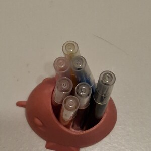 May include: A coral-colored pen holder shaped like a narwhal, filled with various pens. The pens have clear caps and different colored ink, including black, blue, and orange. The holder has a small fin on the side.