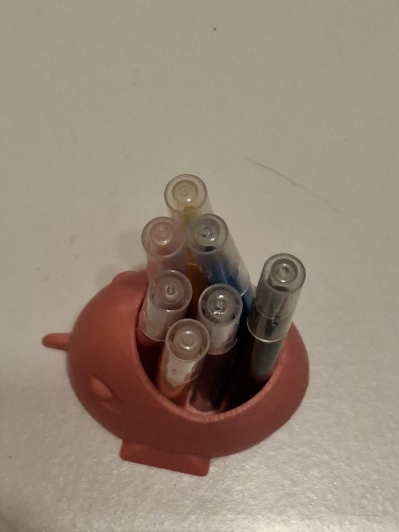 May include: A coral-colored pen holder shaped like a narwhal, filled with various pens. The pens have clear caps and different colored ink, including black, blue, and orange. The holder has a small fin on the side.