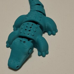 Op de afbeelding: Een turquoise 3D-geprint alligatorfiguur. De alligator heeft een textuuroppervlak, zichtbare poten en een gedetailleerde kop en staart. Het beeldje is een novelty item.