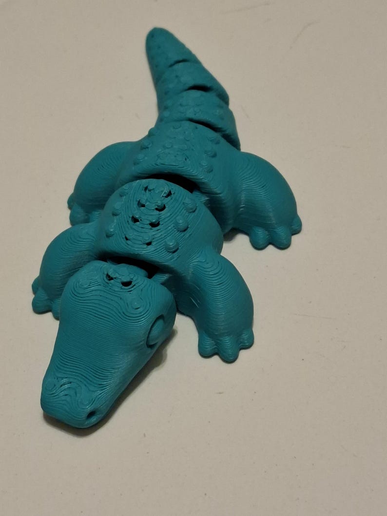 Op de afbeelding: Een turquoise 3D-geprint alligatorfiguur. De alligator heeft een textuuroppervlak, zichtbare poten en een gedetailleerde kop en staart. Het beeldje is een novelty item.