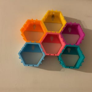 K&ouml;nnte beinhalten: Ein farbenfrohes, wabenf&ouml;rmiges Wandregal. Das Regal besteht aus sechs sechseckigen Regalen in leuchtenden Farben: Orange, Gelb, Rosa, Blau, Lila und T&uuml;rkis. Jedes Regal hat ein Schl&uuml;sselloch zum Aufh&auml;ngen. Die Regale sind an einer wei&szlig;en Wand befestigt.