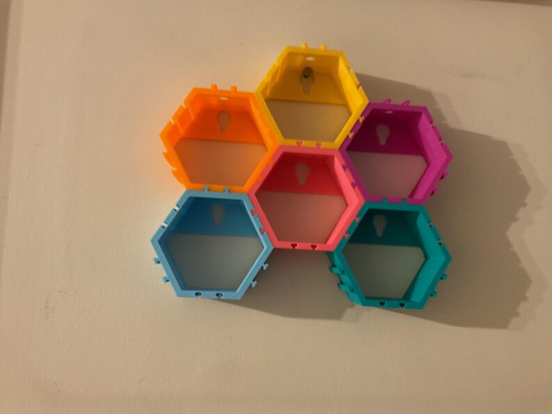 K&ouml;nnte beinhalten: Ein farbenfrohes, wabenf&ouml;rmiges Wandregal. Das Regal besteht aus sechs sechseckigen Regalen in leuchtenden Farben: Orange, Gelb, Rosa, Blau, Lila und T&uuml;rkis. Jedes Regal hat ein Schl&uuml;sselloch zum Aufh&auml;ngen. Die Regale sind an einer wei&szlig;en Wand befestigt.