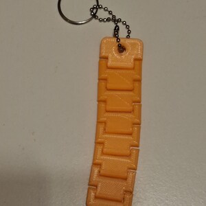 Puede incluir: Llavero de juguete fidget naranja impreso en 3D. El llavero presenta una serie de bloques rectangulares conectados, lo que permite un movimiento flexible. Est&aacute; unido a un llavero de metal y una peque&ntilde;a cadena.