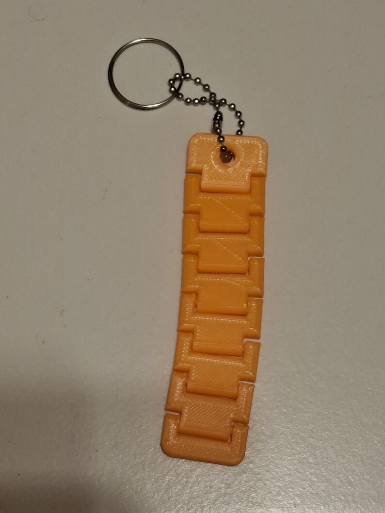 Puede incluir: Llavero de juguete fidget naranja impreso en 3D. El llavero presenta una serie de bloques rectangulares conectados, lo que permite un movimiento flexible. Est&aacute; unido a un llavero de metal y una peque&ntilde;a cadena.