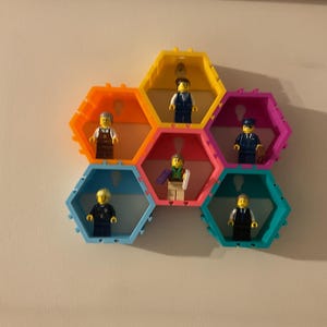 K&ouml;nnte beinhalten: Ein farbenfrohes, sechseckiges Regal mit sechs F&auml;chern, in denen jeweils eine Lego-Figur steht. Das Regal besteht aus hellem Kunststoff in verschiedenen Farben, darunter Orange, Gelb, Rosa und Blau. Die Lego-Figuren tragen unterschiedliche Outfits.