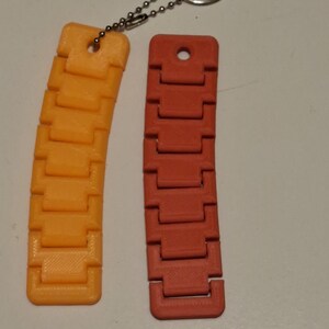 Puede incluir: Dos llaveros segmentados y curvos impresos en 3D. Uno es naranja, el otro es rojo-naranja. Cada llavero tiene una serie de segmentos rectangulares conectados entre s&iacute;. Una cadena y un llavero plateados est&aacute;n unidos al llavero naranja.