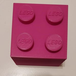 May include: A bright pink LEGO brick with four raised circular studs, each bearing the LEGO logo. The brick is a solid, rectangular shape, and the surface has a slightly textured appearance. The design is reminiscent of classic building toys.