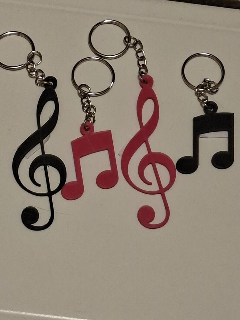May include: Keychains featuring musical notes in black and pink. The keychains have a silver chain and ring. The musical notes include a treble clef and eighth notes. The keychains are made of a solid material.