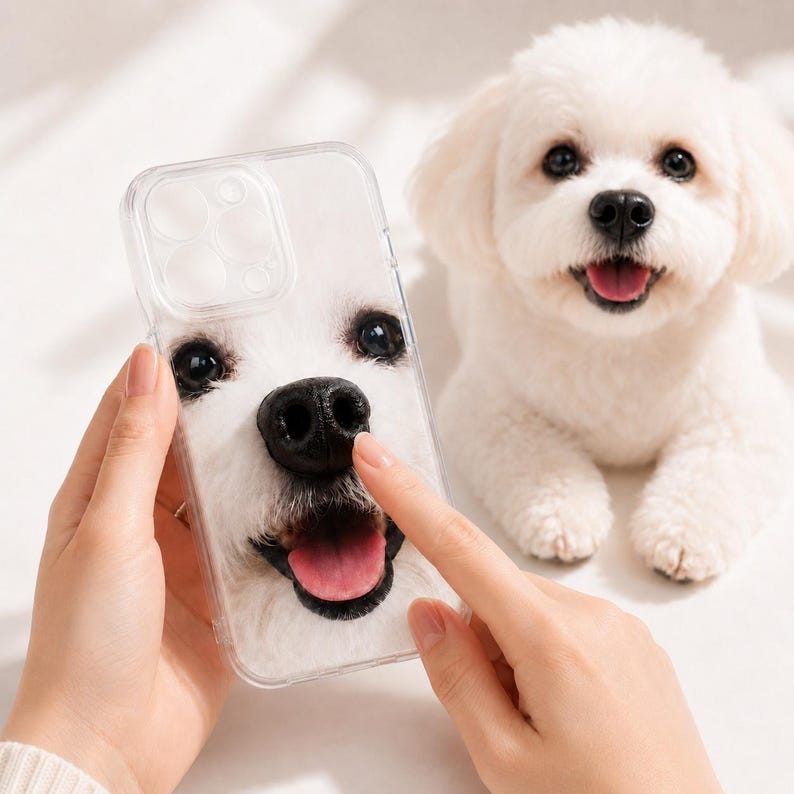 May include: A clear smartphone case featuring a high-resolution print of a white dog's face, including dark eyes, a black nose, and an open mouth with a pink tongue, matching the appearance of the small white dog positioned nearby.