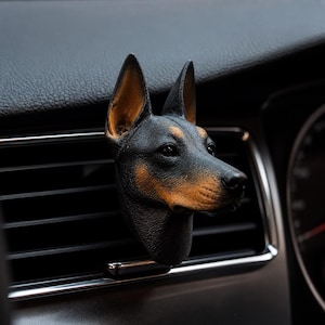 May include: A black and tan dog head car air vent clip. The dog's head is black with tan markings on the snout and ears. The clip is inserted into a car's air vent.