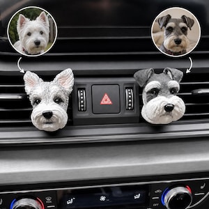 May include: Two dog-shaped car air vent clips attached to a vehicle dashboard. One clip features a white West Highland White Terrier head, and the other features a grey and white Schnauzer head. Both are detailed, three-dimensional sculptures.