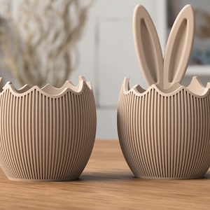 Può includere: Due contenitori beige a forma di uovo di Pasqua con linee verticali. Uno ha orecchie da coniglio. I contenitori hanno un bordo smerlato. Sono su una superficie di legno. Lo sfondo è sfocato.