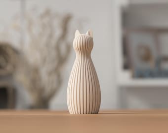 Op de afbeelding: Een lichtbeige, verticaal geribbelde kattenfiguur met een afgerond lichaam en twee puntige oren. Het object heeft een consistente, herhalende lineaire textuur die van de basis tot aan de bovenkant van het hoofd loopt, wat een symmetrische vorm creëert.