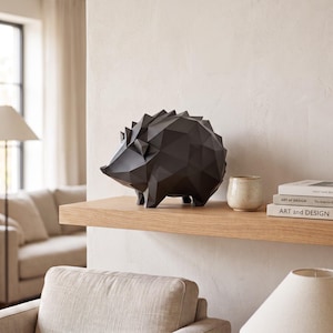 Peut inclure: Une figurine de hérisson noir mat avec un design de surface géométrique à facettes. La sculpture est placée sur une étagère en bois à côté d'une petite tasse en céramique blanc cassé mouchetée et d'une pile de livres avec le texte ART AT DESIGN.