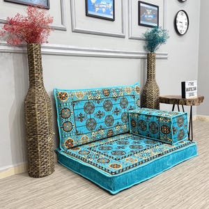 May include: A floor seating set with a turquoise base and patterned cushions. The set includes a large floor cushion, a backrest cushion, and a small ottoman. The cushions feature a detailed design with orange and brown accents. Two decorative vases with dried flowers are on either side.