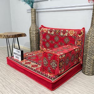 May include: A red floor seating set with a patterned design, including a backrest, a rectangular cushion, and a base. The set is adorned with tassels and is placed on a patterned rug. Two tall decorative vases flank the set, and a small wooden table is to the left.