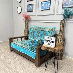 Puede incluir: Un sofá de madera con un cojín y respaldo turquesa y estampado. Un otomán a juego está al lado del sofá. Una pequeña mesa auxiliar con patas de horquilla negras tiene un letrero que dice "THE ANATOLIAN SOUL".