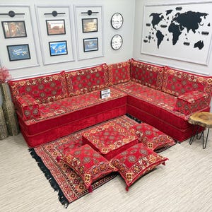 May include: A red sectional sofa with patterned cushions and a matching rug. The sofa is arranged in an L-shape, with several throw pillows and a small wooden side table. The room also features framed artwork and clocks.